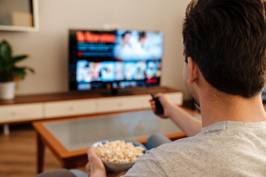 Man watching tv and eating popcorn at home on a couch, back view