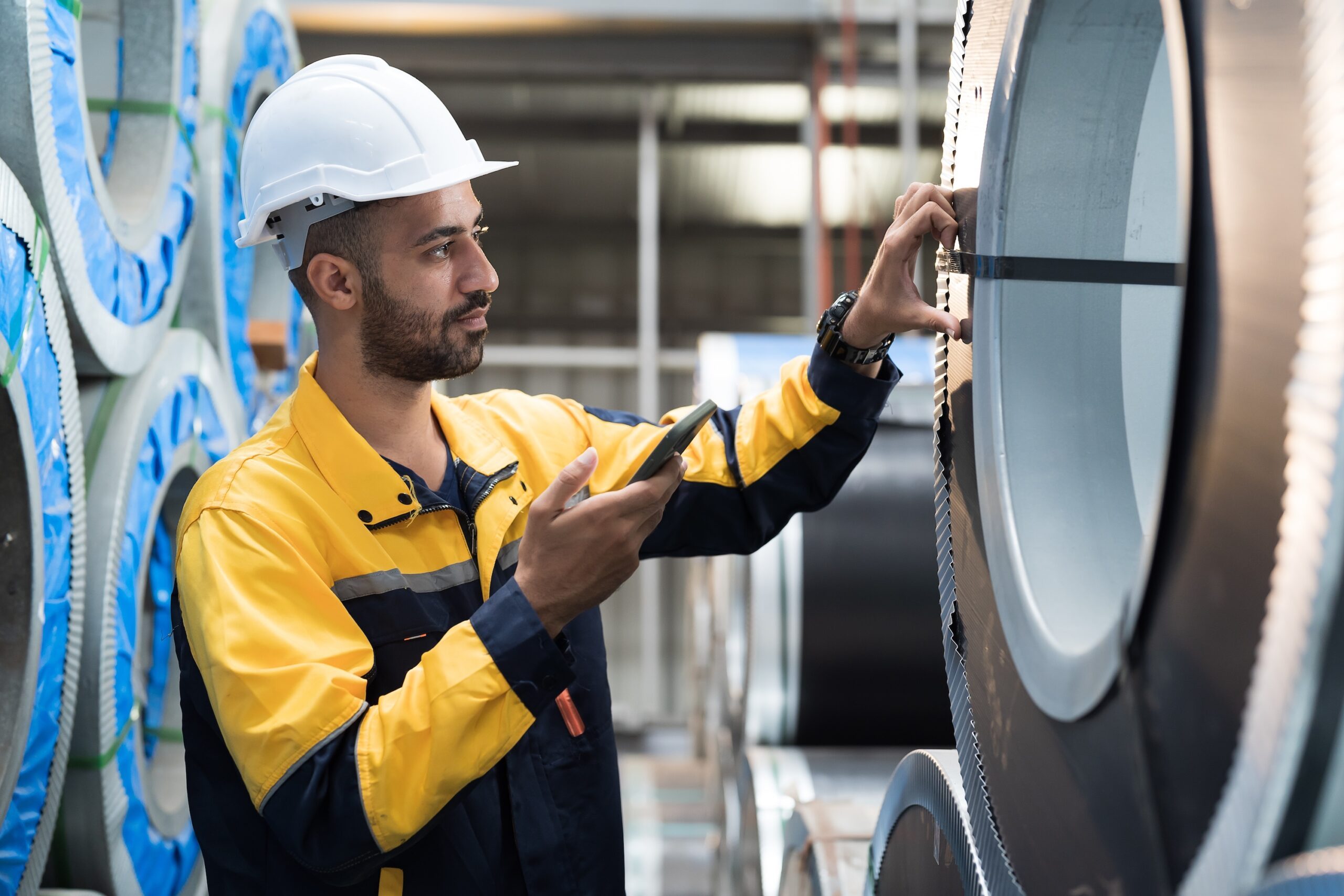 Male factory worker inspecting quality rolls of galvanized or metal sheet in in aluminum material warehouse. Male worker working in warehouse of raw materials during manufacturing process in plant