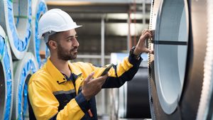 Male factory worker inspecting quality rolls of galvanized or metal sheet in in aluminum material warehouse. Male worker working in warehouse of raw materials during manufacturing process in plant