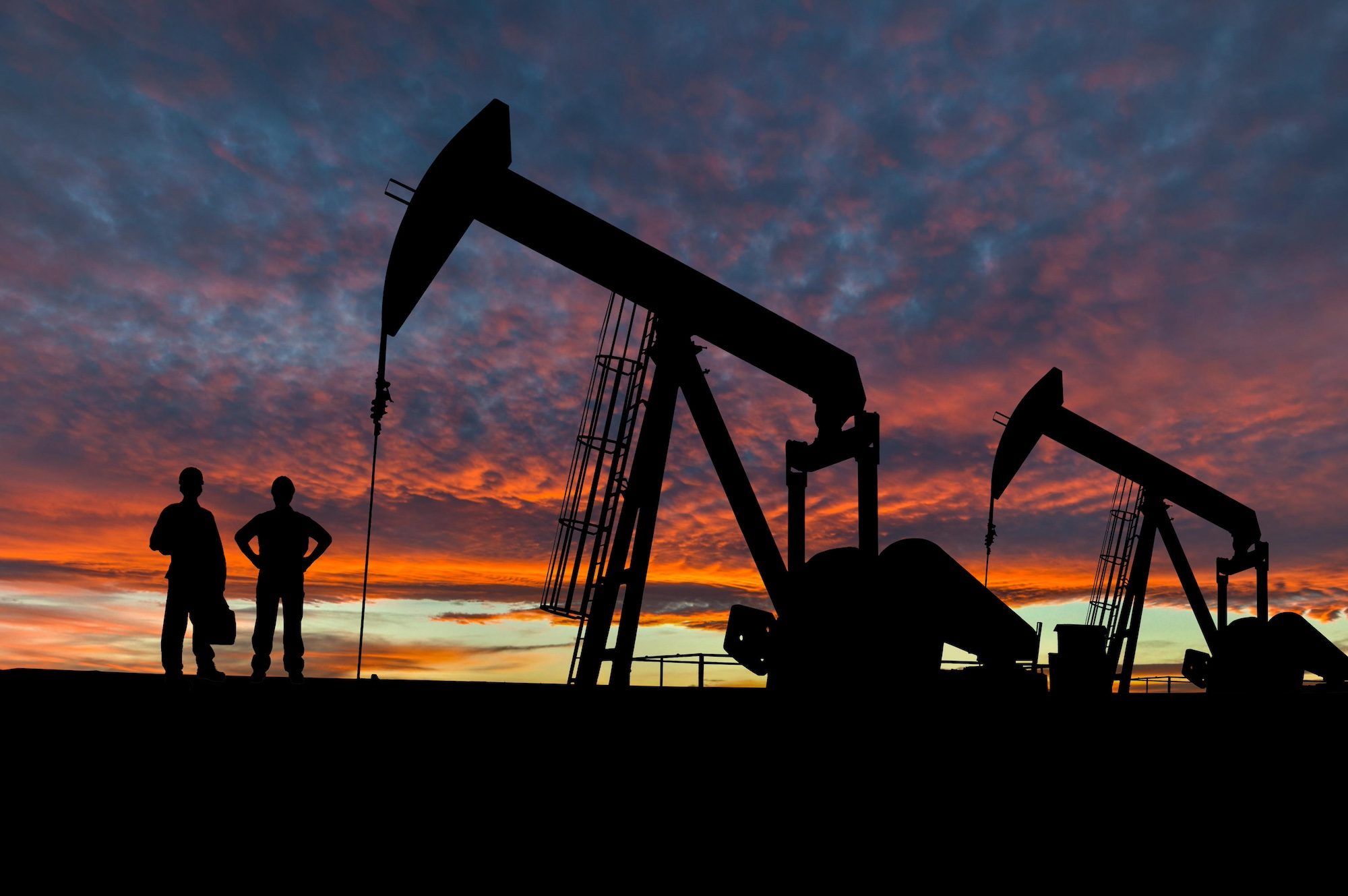 H50RFT Silhouette of oil workers at an oil field pumpjack site against a dramatic sky.