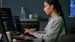 Side view of professional female IT developer from Middle East coding at ease on laptop while sitting at workstation with several computers in office