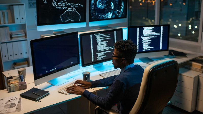 A man sits at multiple computer monitors displaying information. AI can process millions of data points to rapidly generate oil price scenarios