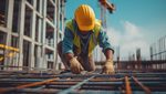 A construction worker wearing a yellow hard hat and safety vest is working on a building site, kneeling on a steel framework. The background shows an unfinished building structure and a crane.