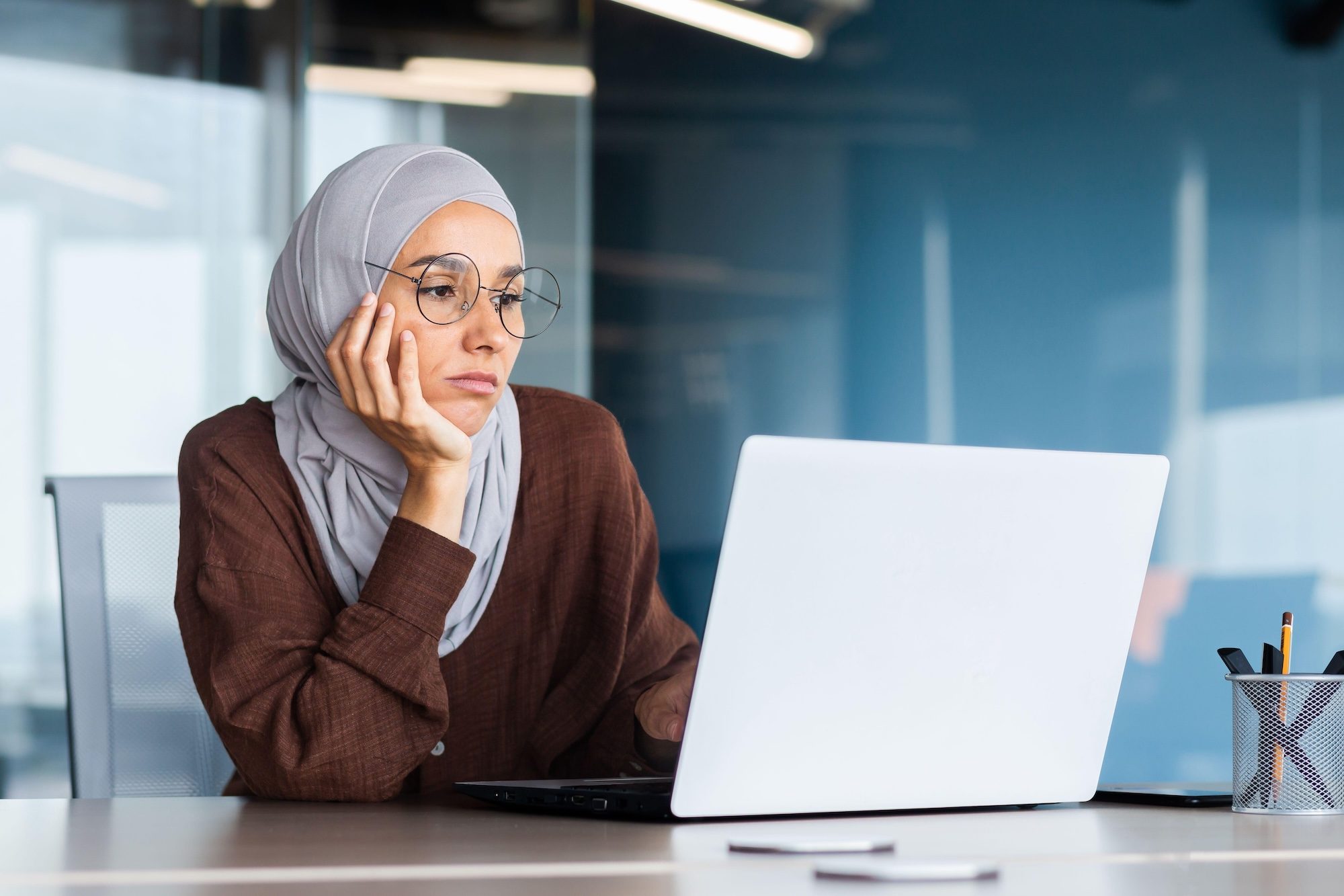 2M92198 Serious bored businesswoman inside office, muslim woman in hijab thinking while sitting at workplace with laptop, woman at work thinking about decisions. female-led, woment-led, female, woman