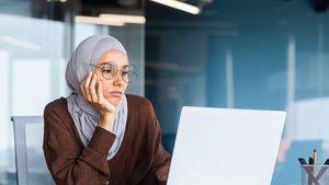 2M92198 Serious bored businesswoman inside office, muslim woman in hijab thinking while sitting at workplace with laptop, woman at work thinking about decisions. female-led, woment-led, female, woman